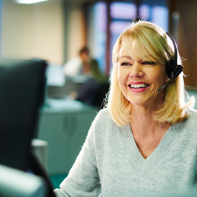 Blonde haired lady sitting at desk in office, wearing a headset and smiling
