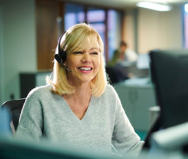 Woman-sitting-in-office-at-pc,-smiling-and-talking-into-headset